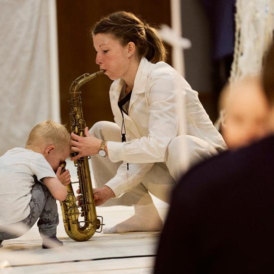 Billedet forestiller en kvinde der sidder på hug med en saxofon. En baby kigger ned i instrumentet.