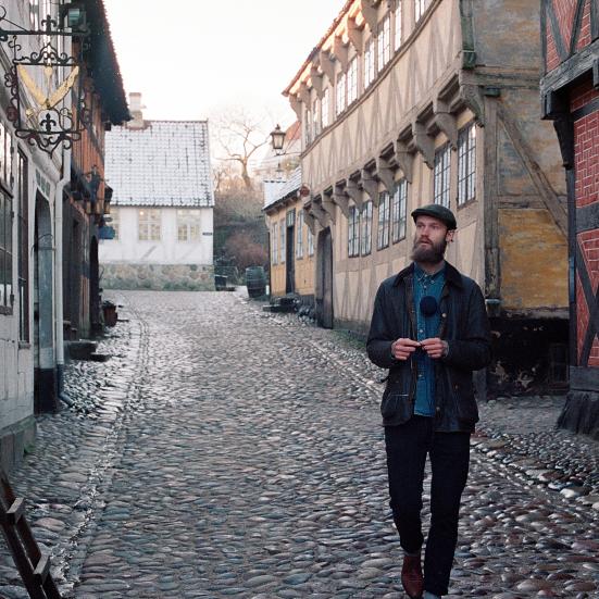 Billedet forestiller den vandrende strikker, der går på en brostenbelagt vej omgivet af gamle huse.