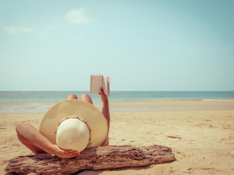 Billedet forestiller en kvinde der ligger på ryggen på stranden. Hun har en lys solhat på og holder en bog op foran sig. Længere væk kan man se havet. 