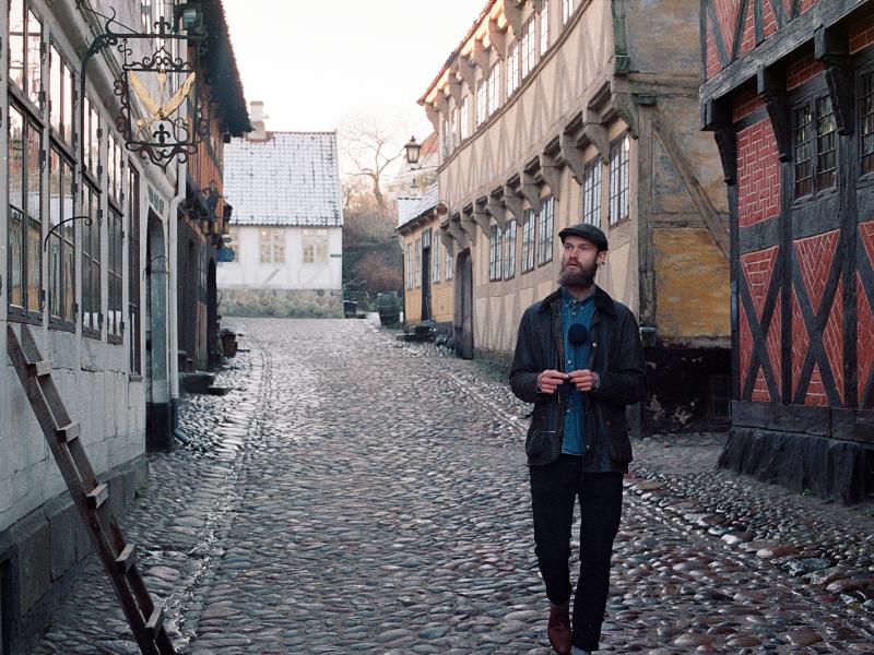 Billedet forestiller den vandrende strikker, der går på en brostenbelagt vej omgivet af gamle huse.