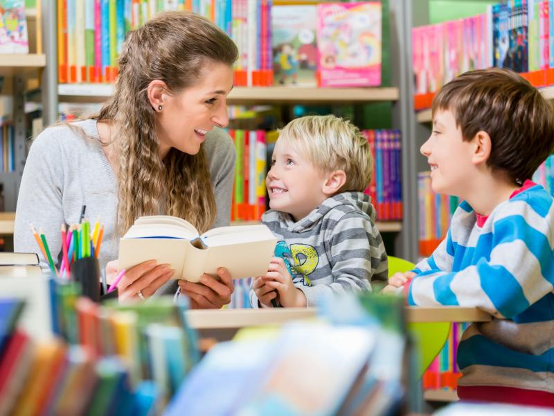 Billedet viser en kvinde, der læser højt for to mindre børn i et bibliotek