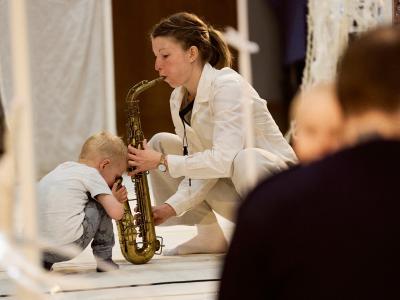 Billedet forestiller en kvinde der sidder på hug med en saxofon. En baby kigger ned i instrumentet.
